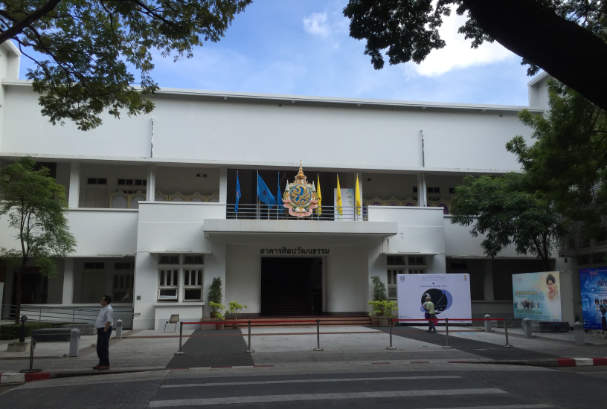 A White two-storey building with flat roof and symmetrical façade. On both floor there are external corridors. The main entrance is marked by a flat canopy and curved side walls. There are flags and an emblem above the entrance canopy.