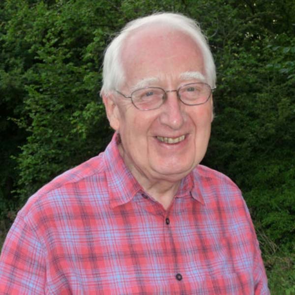 Elderly man with white hair and glasses wearing a pink and blue checked shirt, standing in front of a hedge