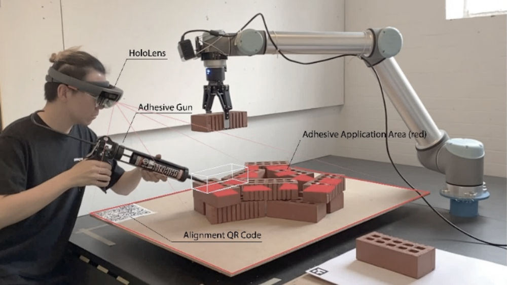 A man wearing virtual reality goggles holds an adhesive gun over a brick structure whist a robot arms holds a brick in place ready to be glued.