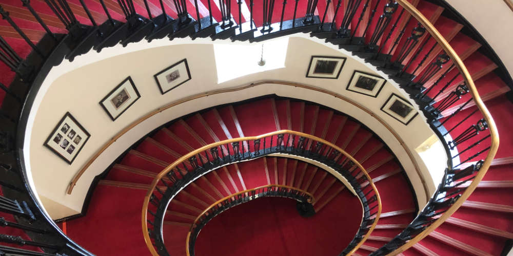 Looking down a Victorian spiral staircase, pictures line the walls.
