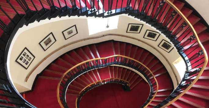 Looking down a Victorian spiral staircase, pictures line the walls.