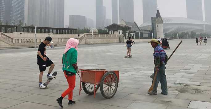 Three people standing on a arge paved square. A young man rests on his skateboard, a woman pushes a wheelbarrow as a man carrying a brush and pan looks on. In the background is a modern high rise cityscape.