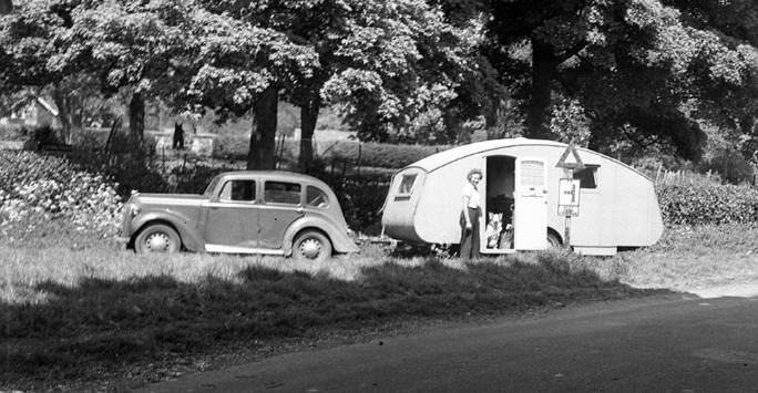 Black and white image of a vintage car and caravan. Photo credit, Museum of English Rural Life, University of Reading pulled over in a lay-by with trees behind. A woman stands by the door and a dog looks out from inside the caravan.