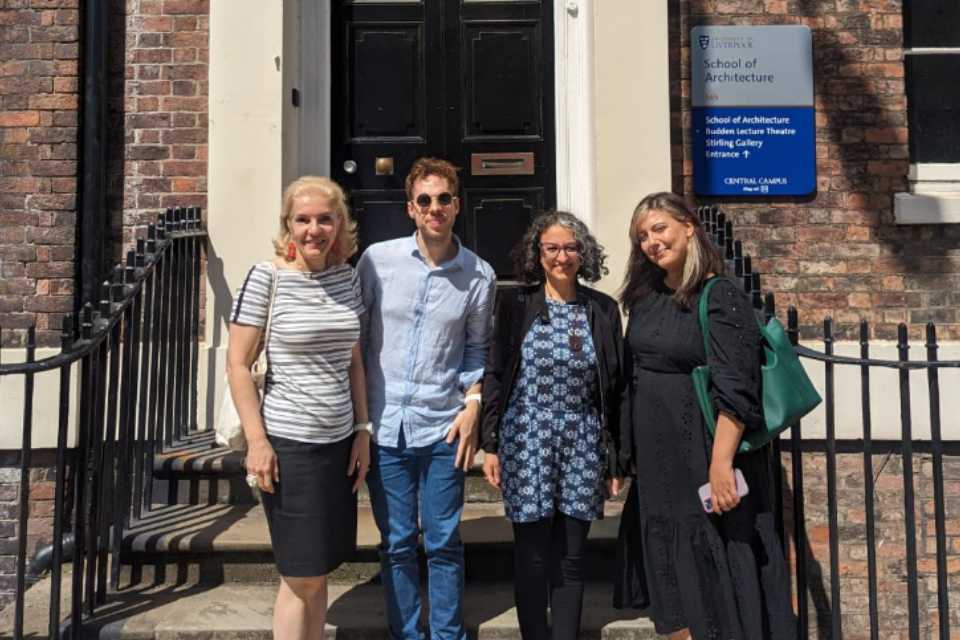 Three women and one man standing on the entrance steps to a georgian building, behind them a black door.
