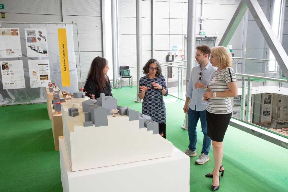 Three women and one man standing around an Architectural model, one of the women is explaining features of the model to the others.