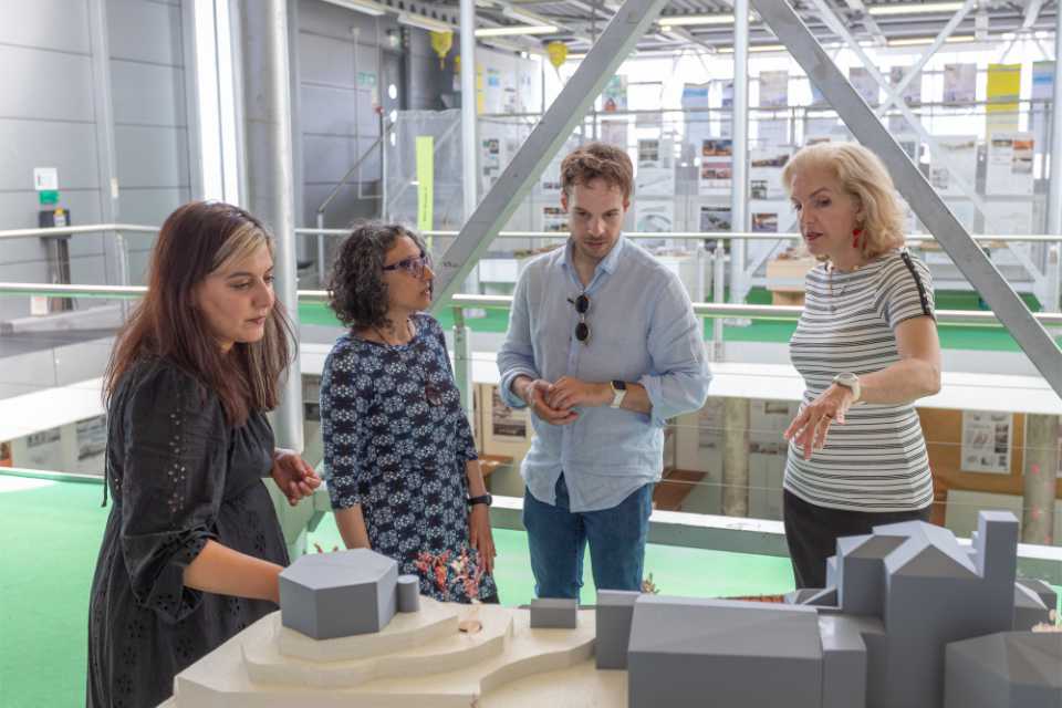 Three women and one man standing around an Architectural model, one of the women is asking a question about the model, pointing to one of the features.