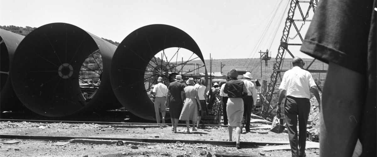 Construction yard with three large steel pipes resting on the ground seen end on. A group of women and men can be seen in ther foreground giving perspective to the huge size.