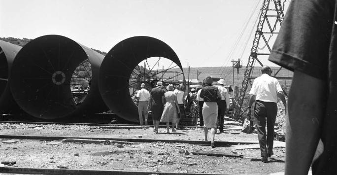 Construction yard with three large steel pipes resting on the ground seen end on. A group of women and men can be seen in ther foreground giving perspective to the huge size.