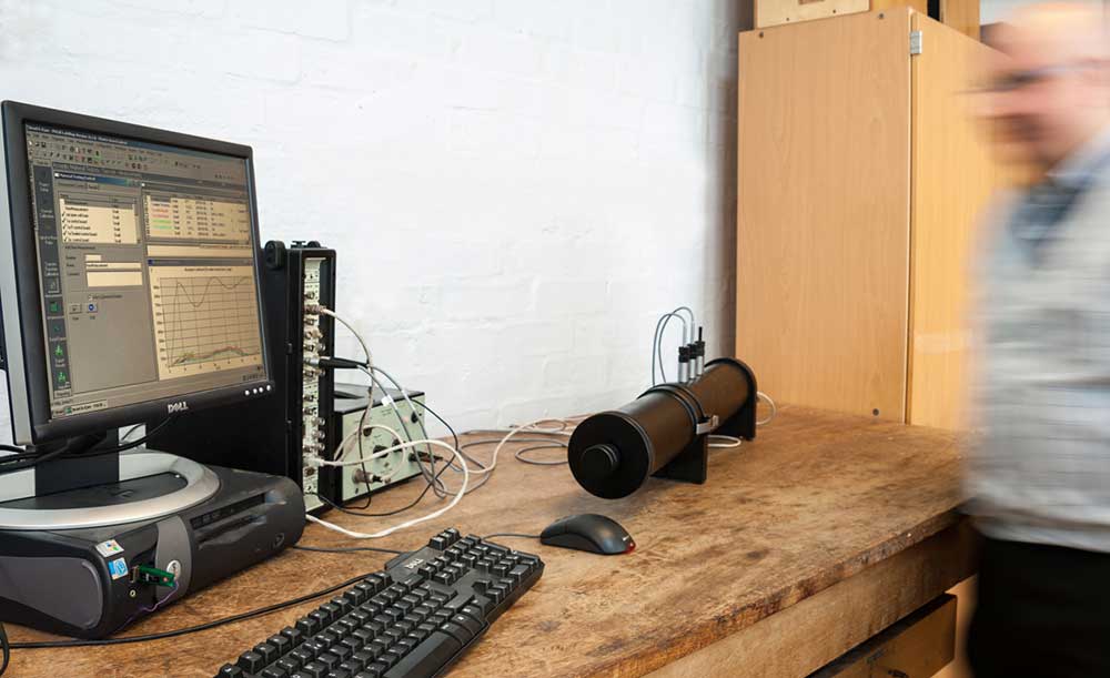 A thick black plastic tube on a wooden bench. Wires coming out of the top are attached to test and computer equipment. A man stands to one side watching.
