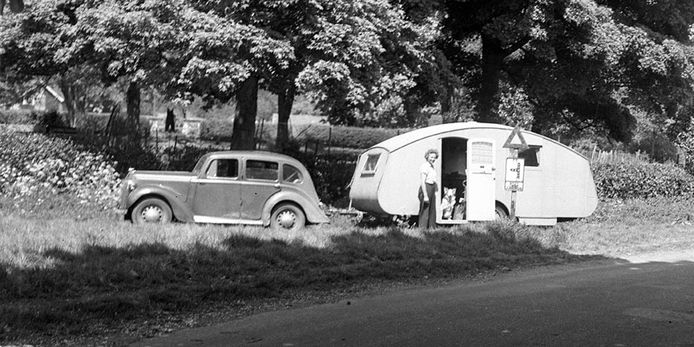 Black and white image of a vintage car and caravan. Photo credit, Museum of English Rural Life, University of Reading pulled over in a lay-by with trees behind. A woman stands by the door and a dog looks out from inside the caravan.