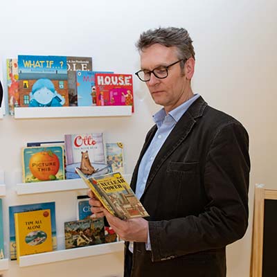 Dr Torsten Schmiedeknecht standing in front of book shelves filled with children's books, reading a Ladybird book on Nuclear Power
