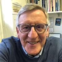 Middle aged man smiling at the camera. He has greying hair, is wearing glasses, a shirt and jumper.