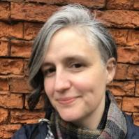 Headshot of a Woman with an asymmetric bob haircut and wearing a scarf and jacket standing in front of a brick wall.