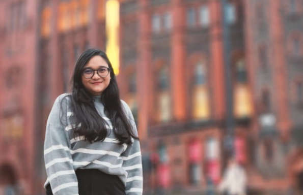 Woman with long hair standing in front of a victorian brick built building. She is smiling and wearing a stripy jumper.