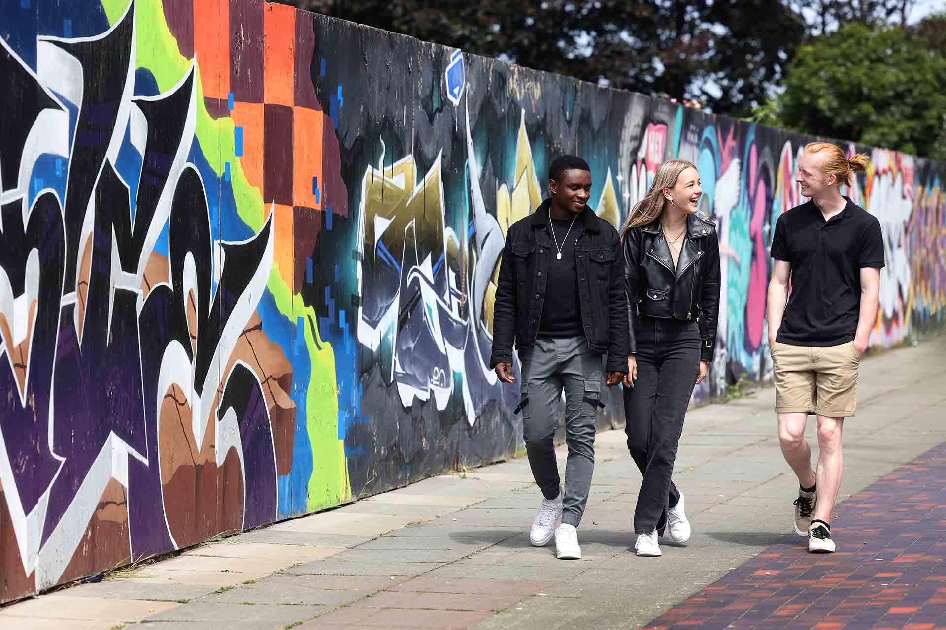 Three students walking next to a wall of colourful artwork