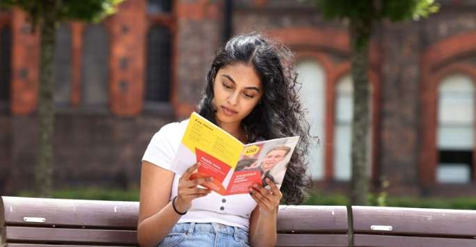 A student sits on a bench and reads an information booklet on the University of Liverpool campus