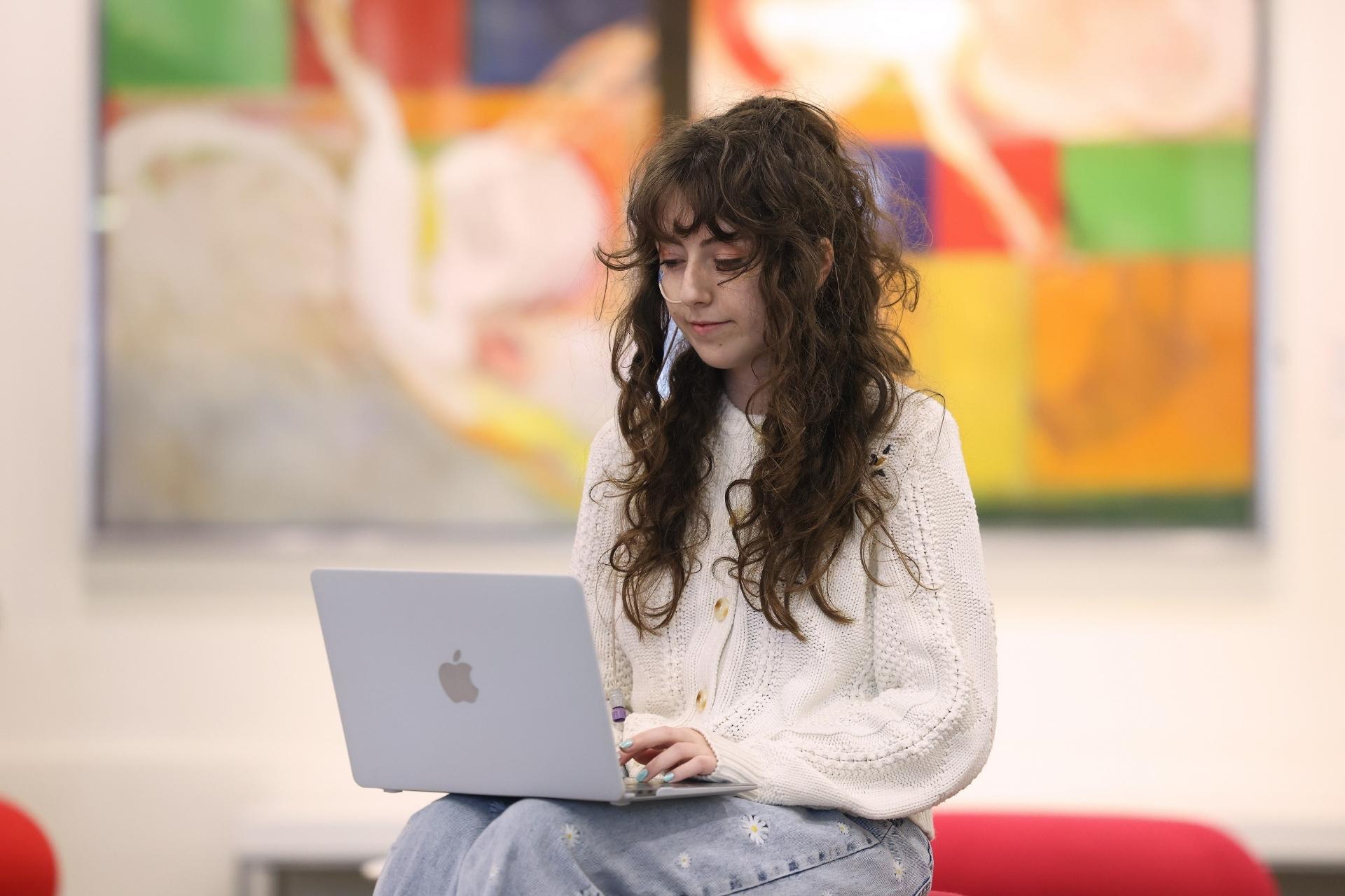 A student sitting confidently with a laptop, focused on their work in a modern, inclusive space with vibrant abstract art in the background.