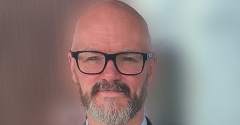 Neill is smiling into the camera. He is wearing glasses and has a neat close shaven beard.