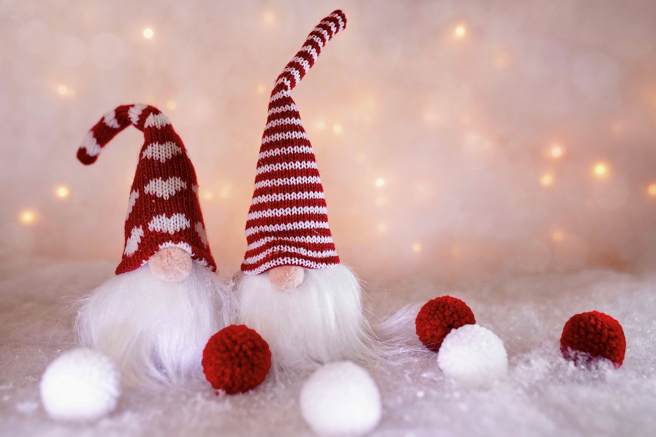 Two gnome toys with red and white hats on, in front of a snowy backdrop with yellow string lights