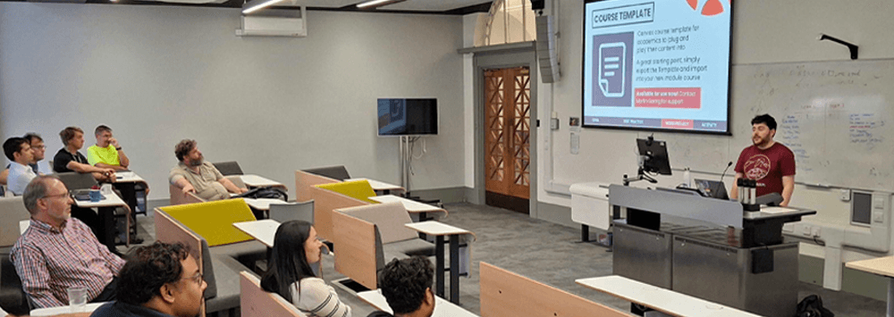 A view from the back of a university lecture theatre with James stood at the front, presenting a workshop in front of large screens to a group of a dozen attendees.