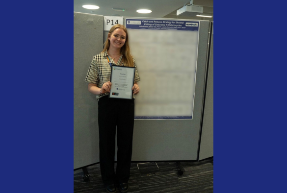 A woman stands with a certificate, smiling in front of a large poster showcasing an event theme.