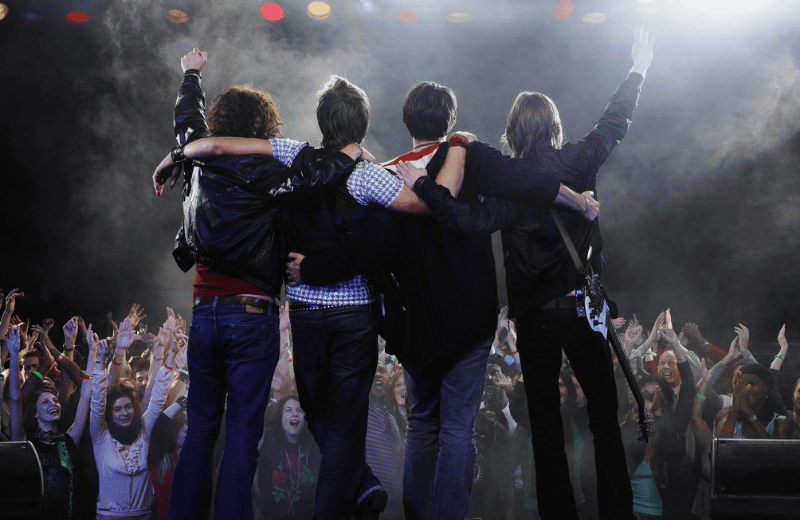A band standing on stage look out to the crowd, who are waving their arms in the air
