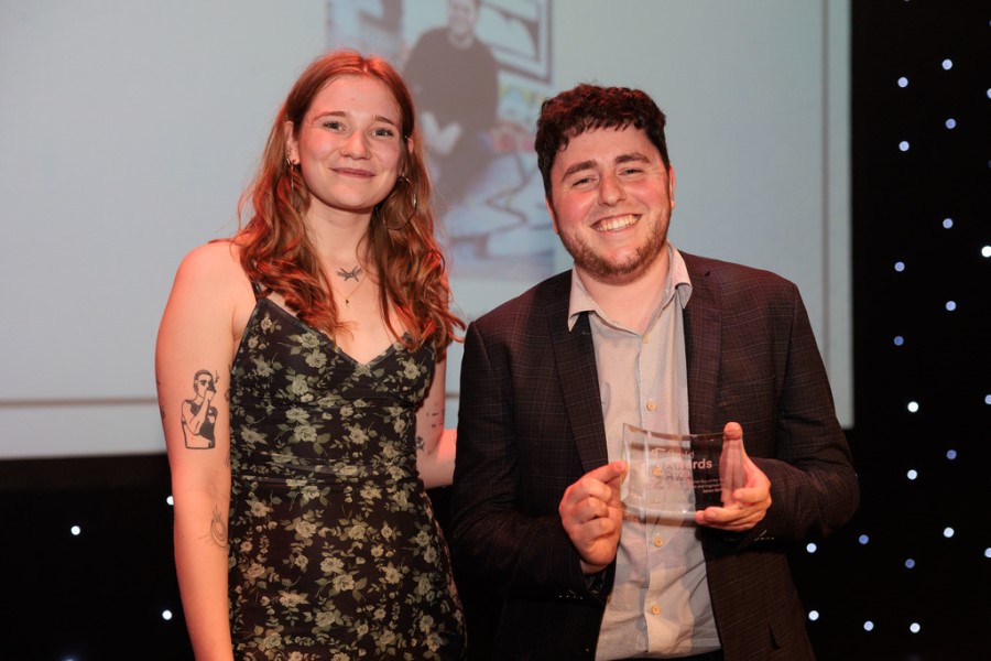 A smiling man in a dark suit with a white shirt holding a glass award and a smiling lady with long hair in a black floral dress