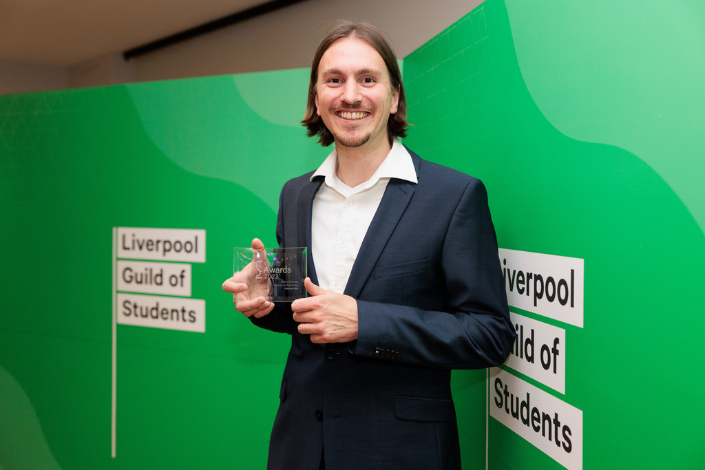 A smiling man in a dark suit with a white shirt holding a glass award