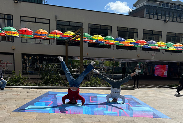 two students doing headstands on an optical illusion installation on University Square