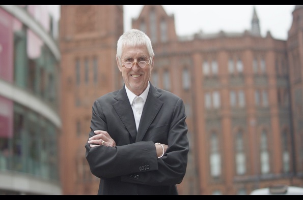 A male Professor stood in front of a red brick building