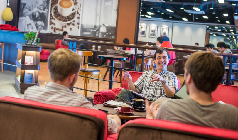 Three students sitting in a cafe with more students in view behind them