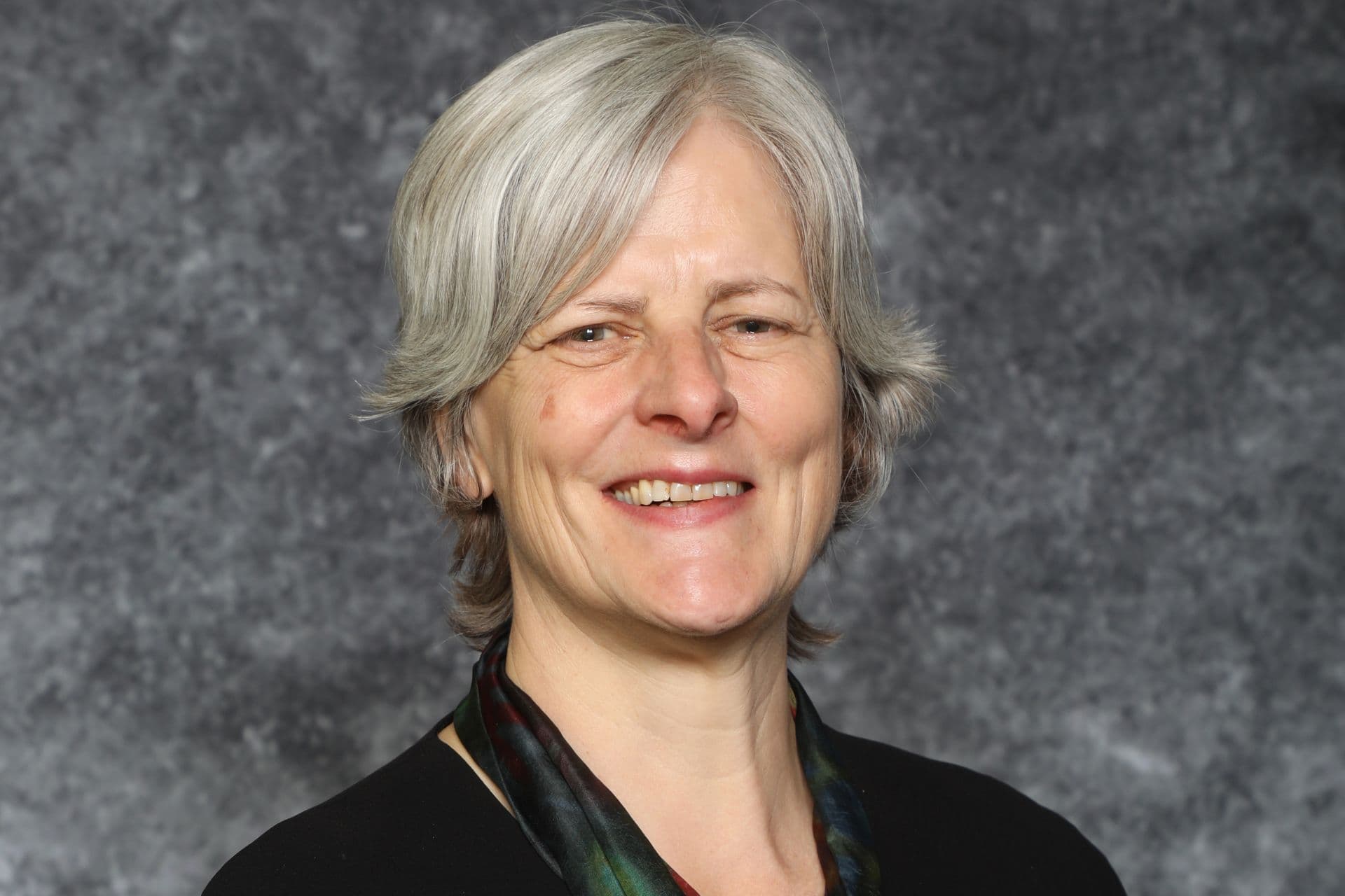 Fiona is smiling, has short, grey hair and a black jacket with a silk scarf tied loosely around her neck.