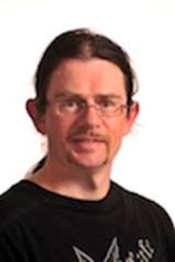 An image of a white male with thin framed glasses on, with long brown hair tied back. He is wearing a black t-shirt.