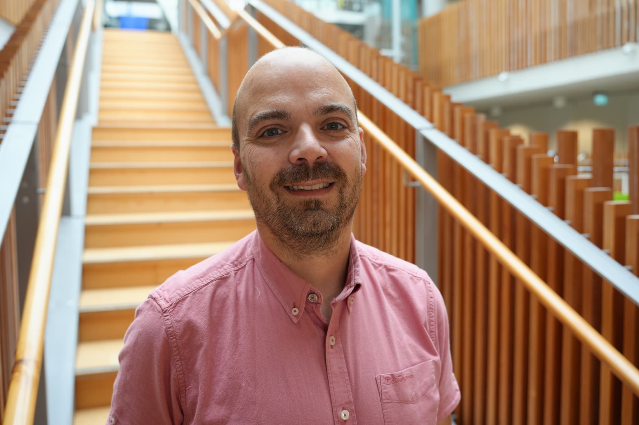 A smiling man wearing a pink shirt stands in front of a wooden staircase inside a modern building.