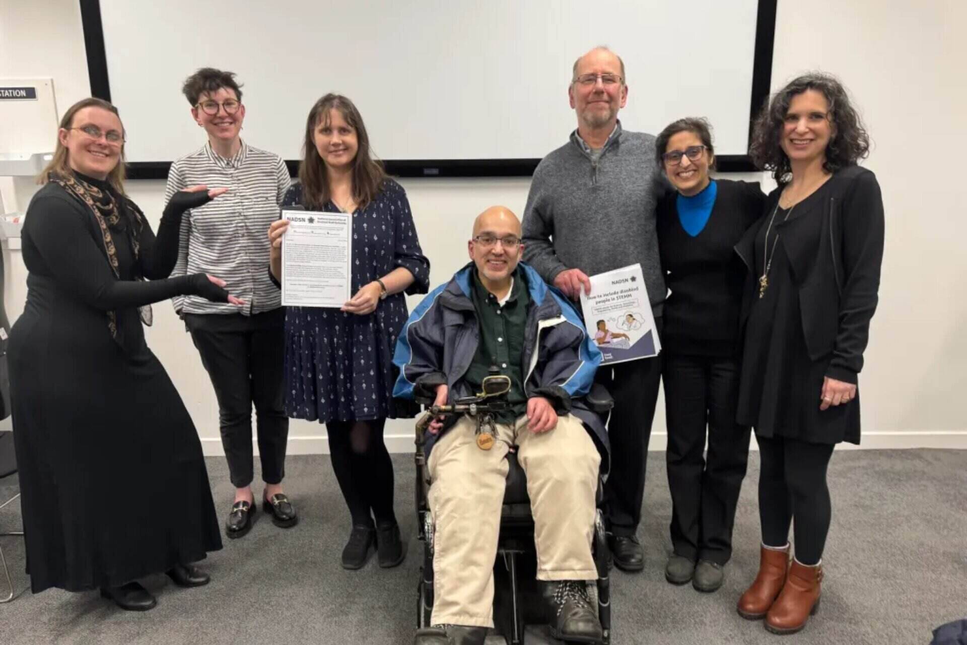 Seven people, including one person using a wheelchair, smile and hold the NADSN STEMM Action Group White Paper. They are in a bright classroom setting with a large screen behind them.