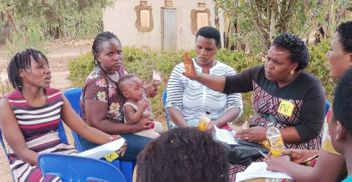 Group of women seated in a semi-circle having a discussion outdoors, with some holding refreshments and one woman gesturing while speaking