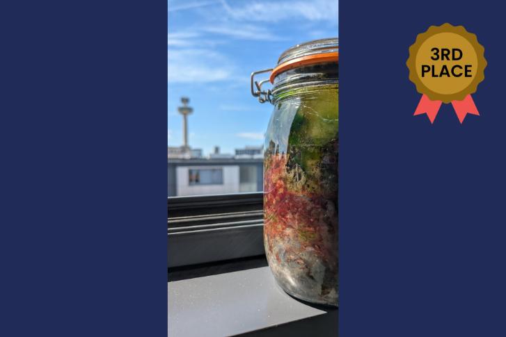 A picture of a jar containing water from the River Mersey, egg yolk, and newsparer on a window ledge overlooking Liverpool against a navy blue background with a bronze 3rd place in the top right corner