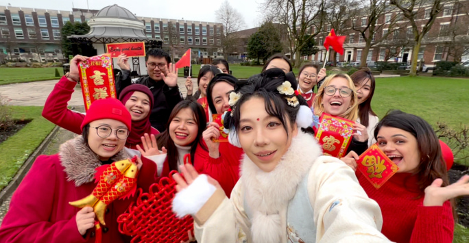 A group of international students wearing red and white and holding Lunar New Year ornaments in Abercromby Square. They are all waving and smiling at the camera.