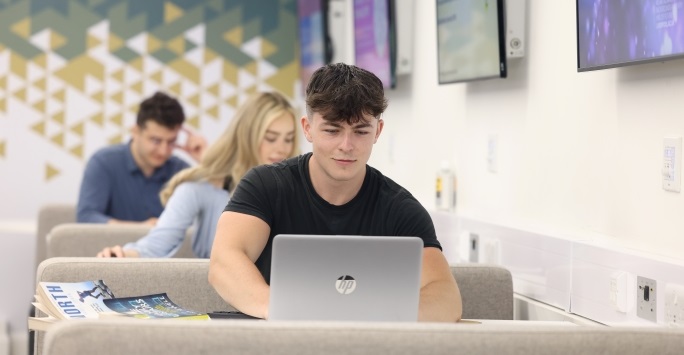 A male student smiles while using a laptop in a university classroom