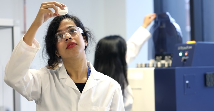 A student wearing safety goggles and a white lab coat holds up a lab sample