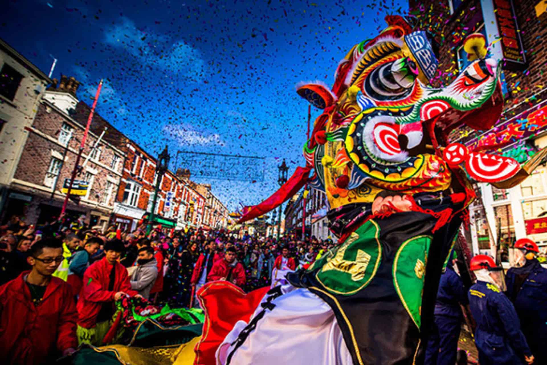A crowded street in Liverpool is full of people celebrating Chinese New Year. There is a large, colourful lion dance costume in the foreground and confetti in the air.