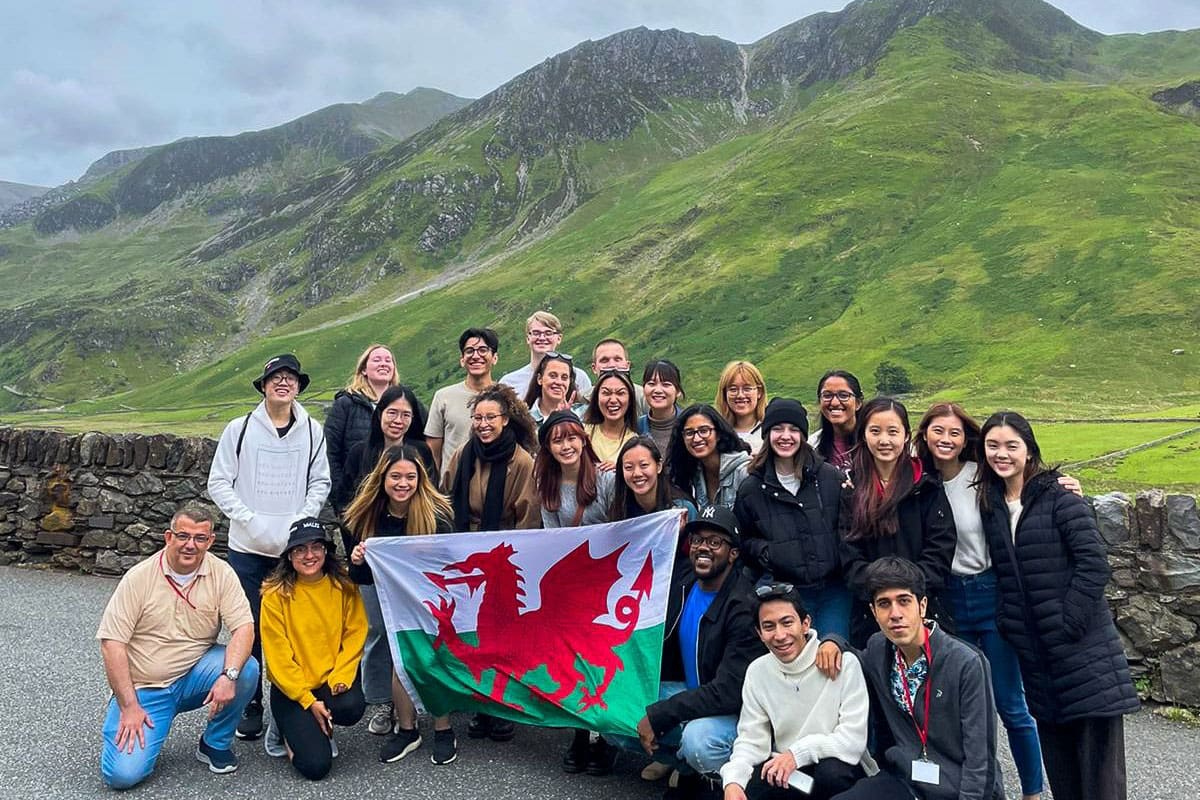 A large group of students in front of a green hill. They hold a Welsh flag.