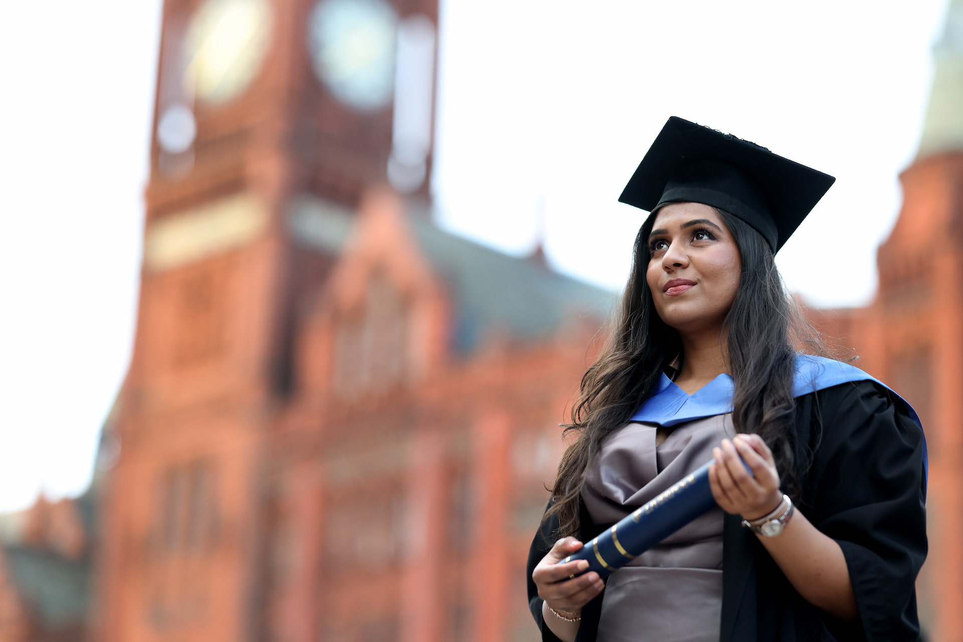 A student stands proudly with her degree in front of the red brick Victoria Building
