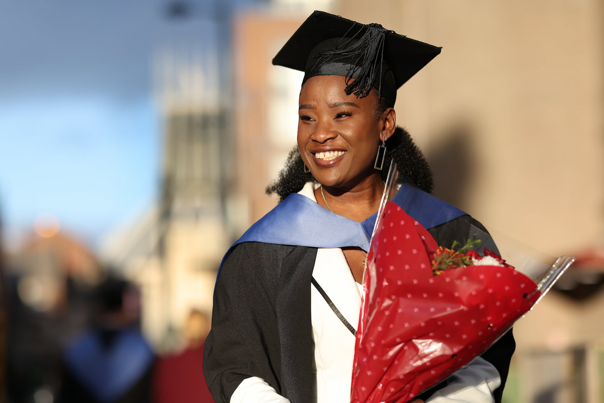 A student standing in the sun holding a bouquet of flowers on graduation day