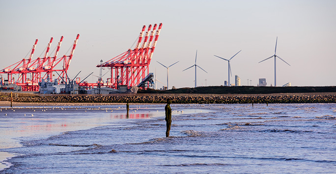 Iron sculpture of a man standing in the waves looking out to sea with dock cranes in the background