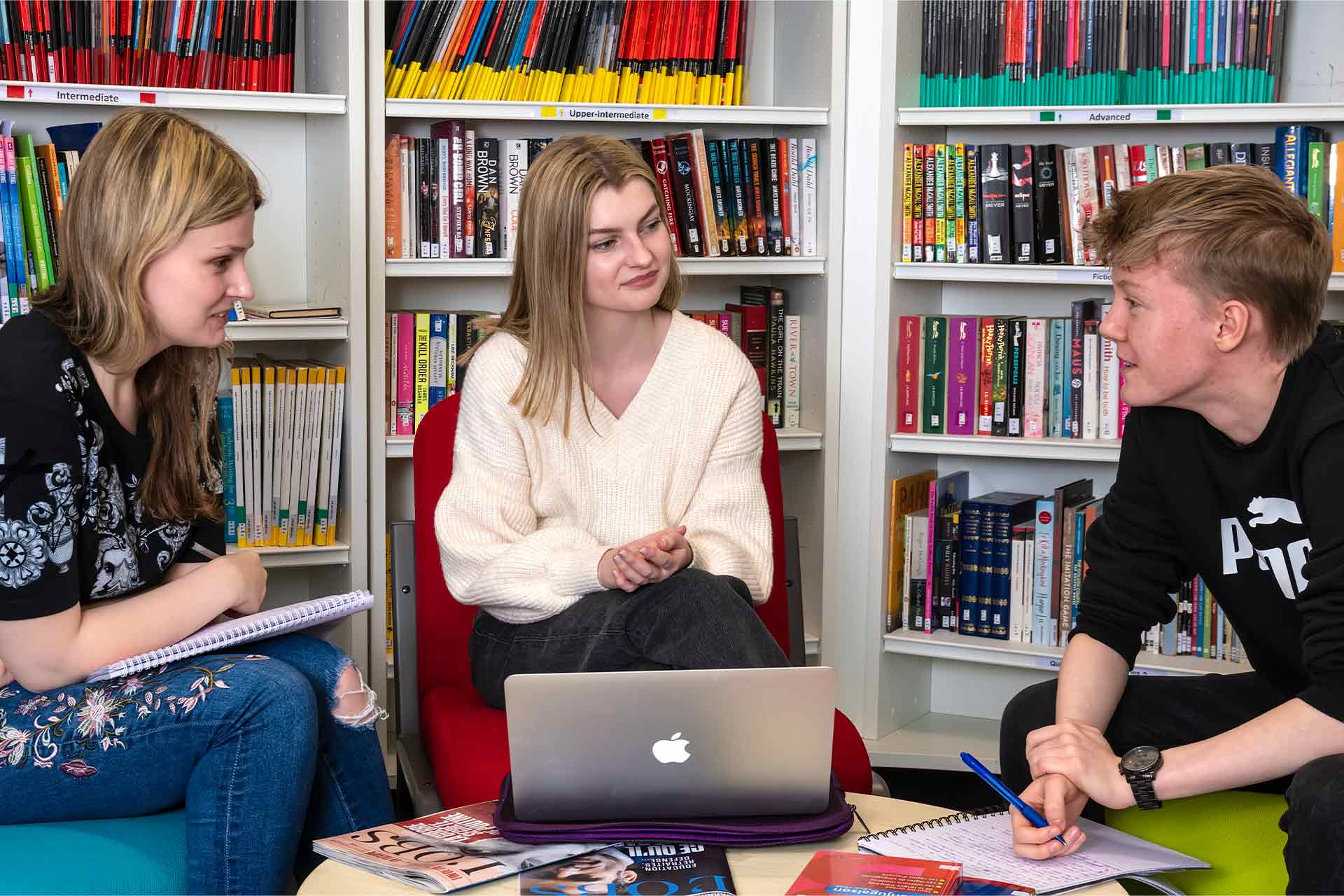 Three students sitting in a library at a table with a laptop and books in discussion with each other.