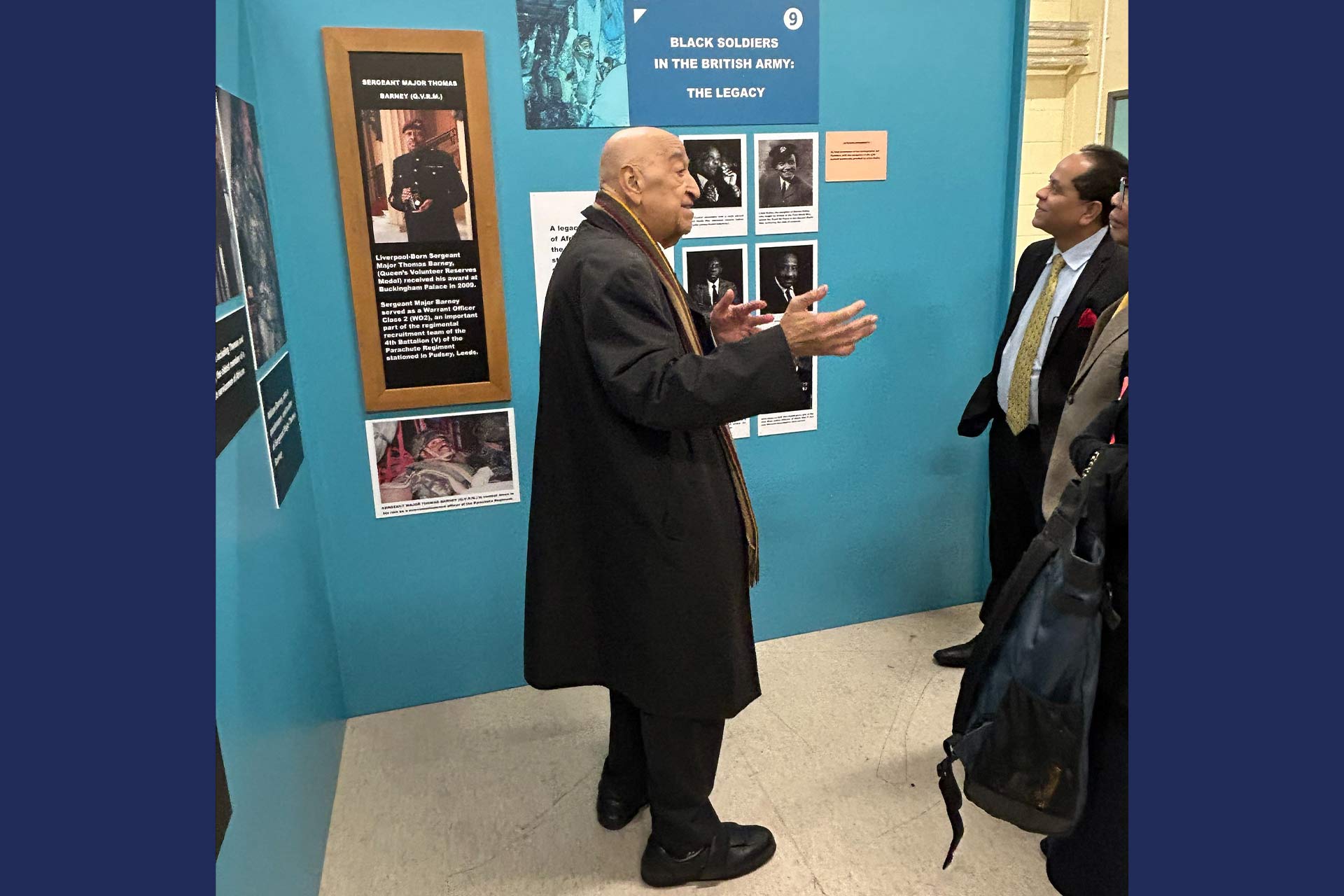 An image of a man in a museum gesticulating and talking to a group of people, in the background some of the material reads 'Black Soldiers in the British Army'
