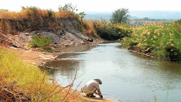 a man crouching down with his back to camera in front of a river, looking in the river bank