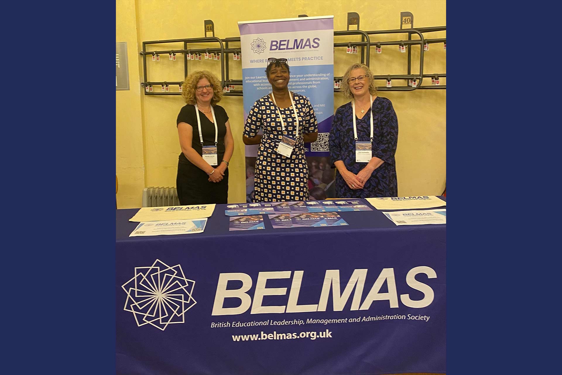 Three women standing behind a desk at a conference, with marketing material reading 'BELMAS - British Educational Leadership, Management and Administration Society'
