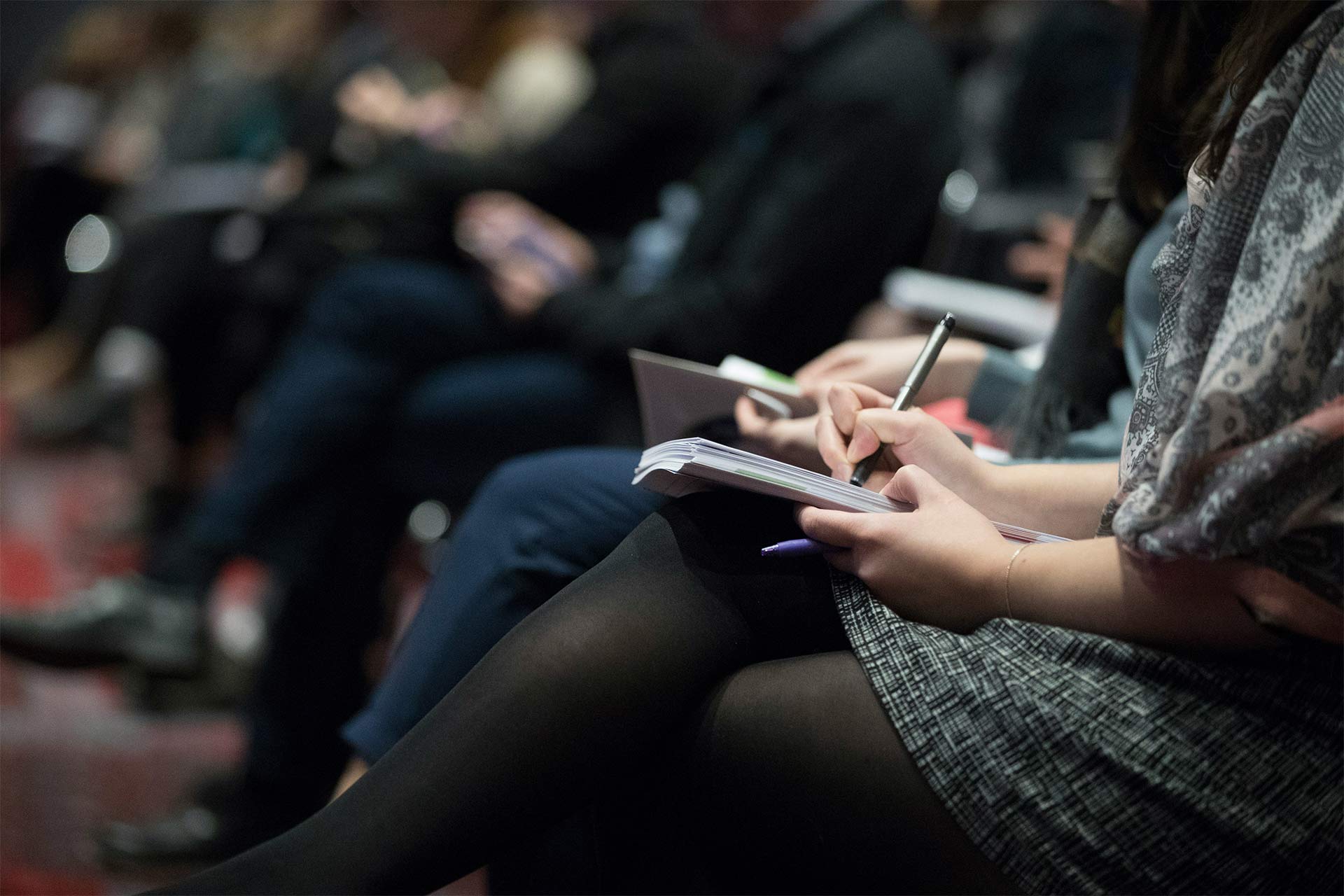 People sitting in a room attending a conference or seminar, a person at the front writes notes with a pen and paper.